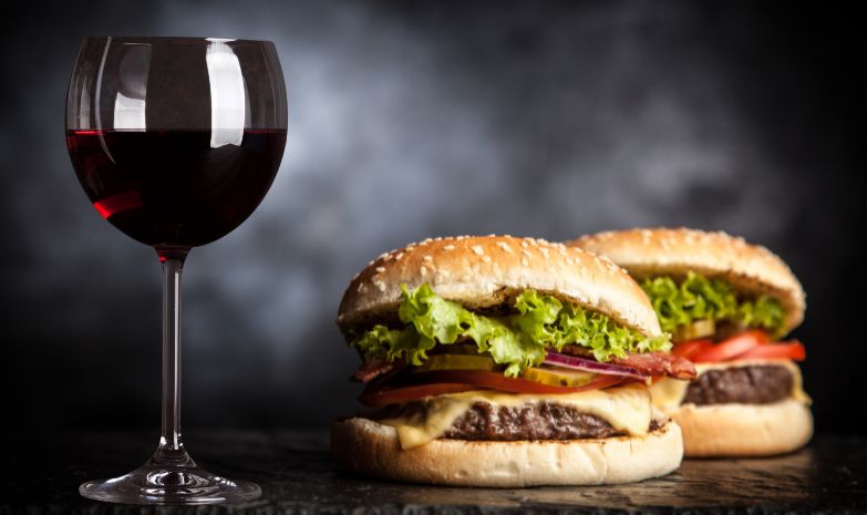 Das Food-Pairing-Konzept kombiniert ein elegantes Glas Rotwein mit zwei herzhaften Cheeseburgern auf einem dunklen Tisch.